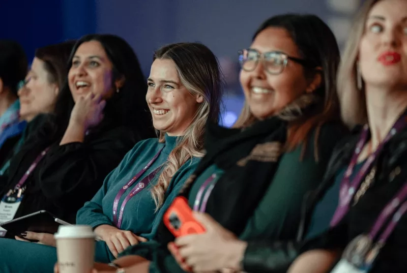 Five women sitting at a meeting, smiling and holding a tablet, cell phone, and coffee cup.