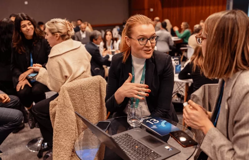 Two women engaged in conversation at a conference with other people in the background.