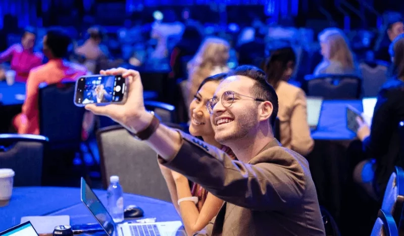 A man and woman taking a selfie at a conference with other people in the background.