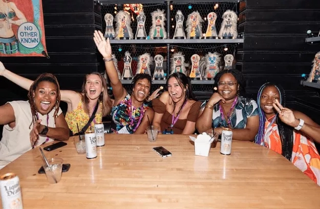 Group of smiling women sitting at table with cans of Liquid Death in front of shelves of figurines.