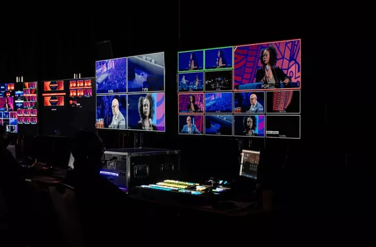 Several monitors and screens show different people talking in a virtual town hall meeting.