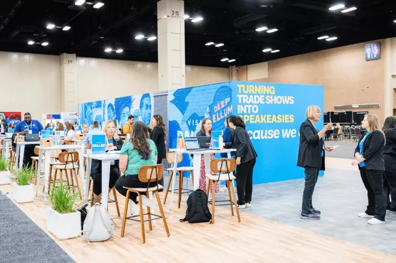 A group of people are sitting and standing at tables with a large blue sign in the background.