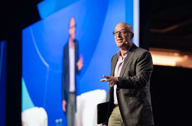 A man is standing on stage in front of a screen, speaking into a microphone, and gesturing.