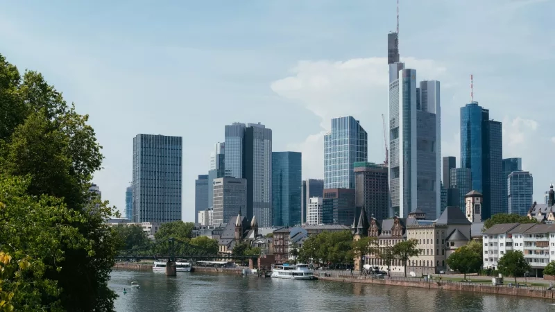 The skyline of Frankfurt, Germany, features high-rise buildings and a river with boats.