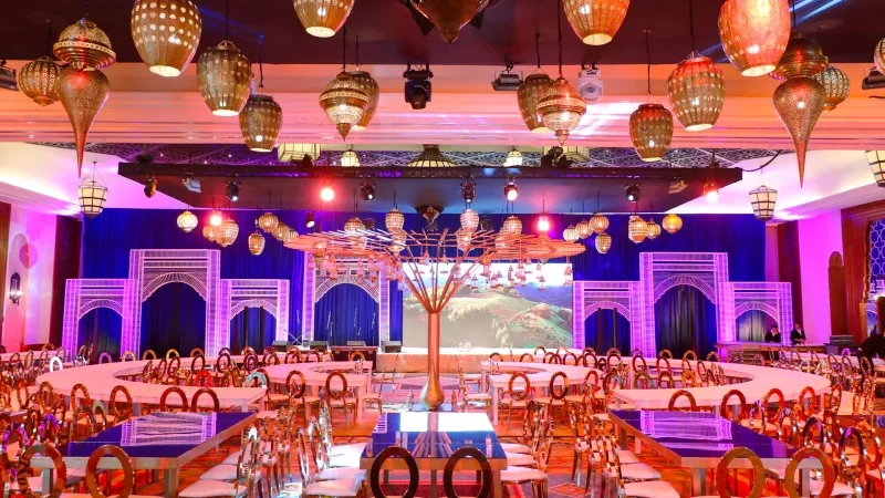 A large banquet hall with tables, chairs, and a stage with a large screen and lanterns.