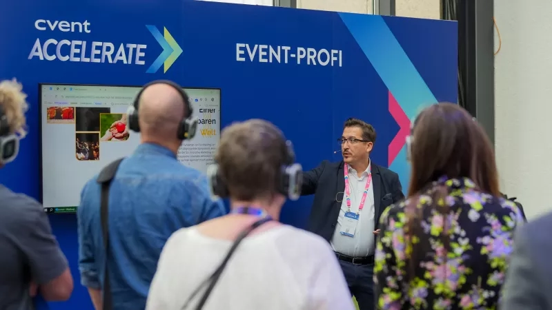 Group of people at Cvent Accelerate conference, standing around a large monitor with headphones on.