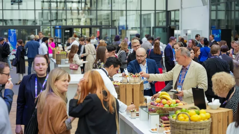 A group of people at a conference, some standing at a table with fruit and drinks.
