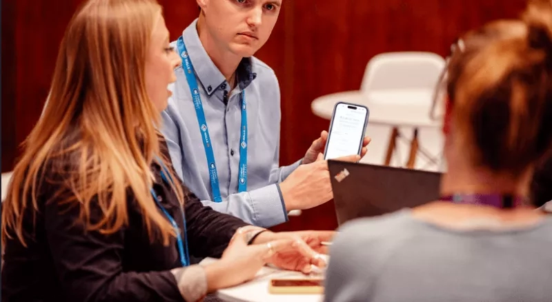 A man is showing a woman a smartphone while she is holding a laptop on the table.