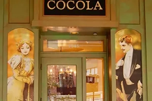 Front entrance of Cocola chocolate shop with green walls and a yellow-dressed woman painted on the left wall.