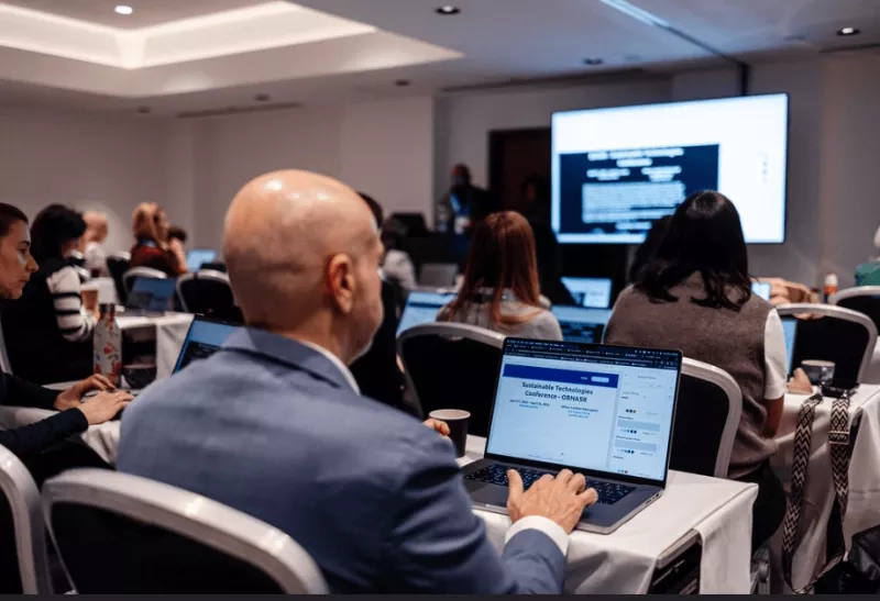 A group of people sitting at tables with laptops, a large screen in the background, and text on the laptop.