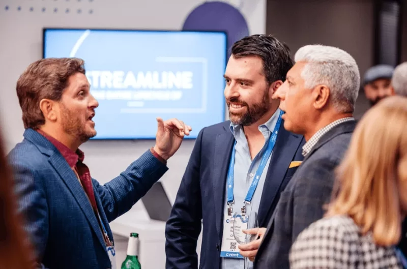 Three men and a woman chatting in a conference room, with a large screen showing the word "STREAMLINE".