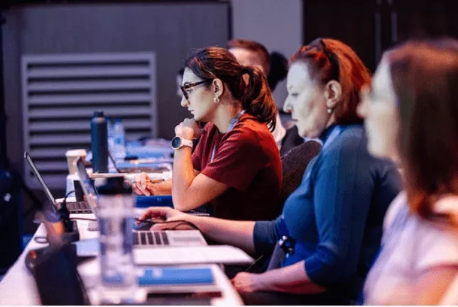 Several women are working in a conference room with laptops, water bottles, and a charger on the desk.