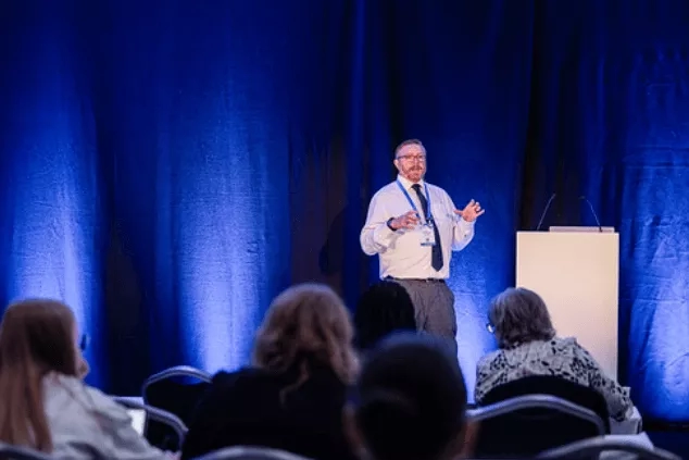 Man standing on a stage giving a presentation to a group of people in chairs.