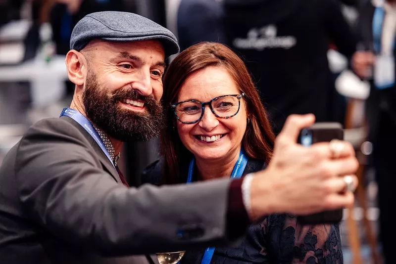 A man and a woman are smiling while the woman is taking a picture of them with her cellphone.