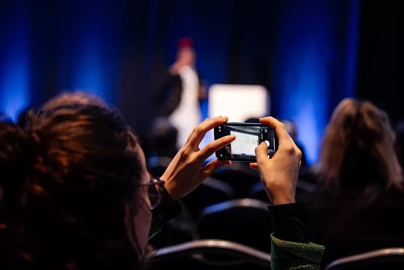 A woman is taking a picture of a person on a stage with a cell phone.