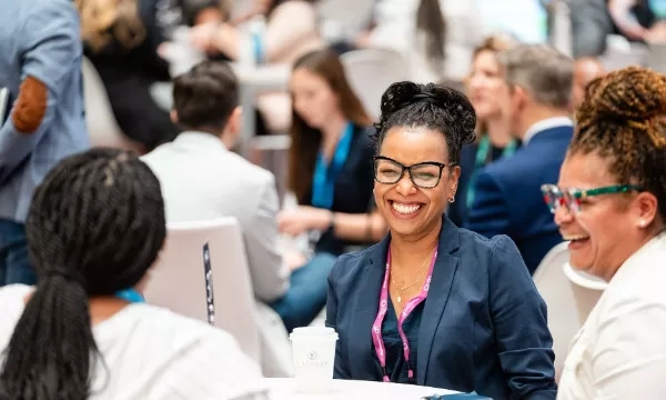 Woman in glasses and blazer smiling at a conference with others in the background.