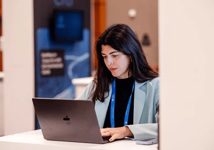 Woman working on laptop at a table with a banner on the wall behind her