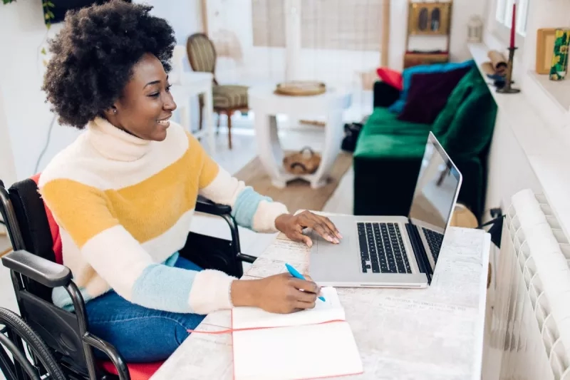 A woman in a wheelchair uses a laptop and pen to take notes in her home office.