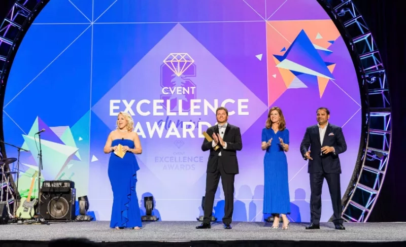 Four people on stage with microphone and trophy for Cvent Excellence Award with purple background and geometric shapes.