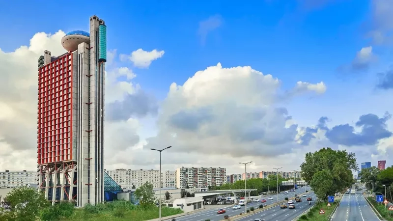 Hyatt Regency Barcelona Tower with highway and buildings in the background under a cloudy sky.
