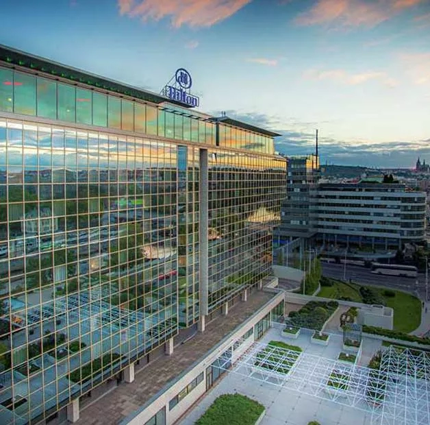 A top view of the Hilton Prague, showcasing its glass facade, logo, and surrounding urban landscape at dusk.