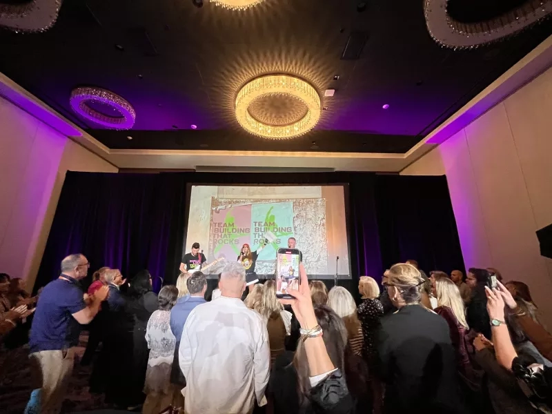 A group of people are standing and raising their hands in a hall with a projector screen.