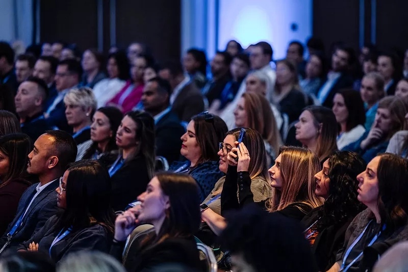 A crowd of people is sitting in a conference hall and listening to the speaker, with blue lights on.