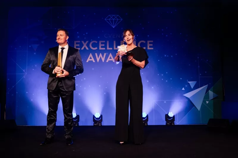 A man and a woman standing on stage holding trophies and a flower in their hands.