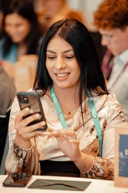 Woman with a badge around her neck looking at her phone at an event with others in the background.