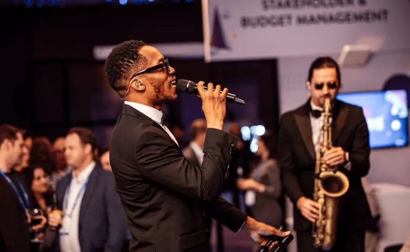 A man sings at an event while another plays the saxophone, and people mill about.