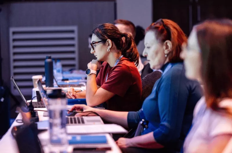 A group of people are sitting in front of a table with laptops and bottles on it.