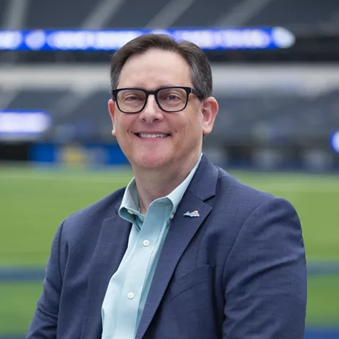 Adam Burke wearing a suit and smiling in front of a stadium with a field and empty stands.