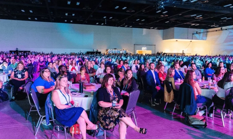 A large conference room with many people seated on chairs at the tables, with purple and blue lighting.