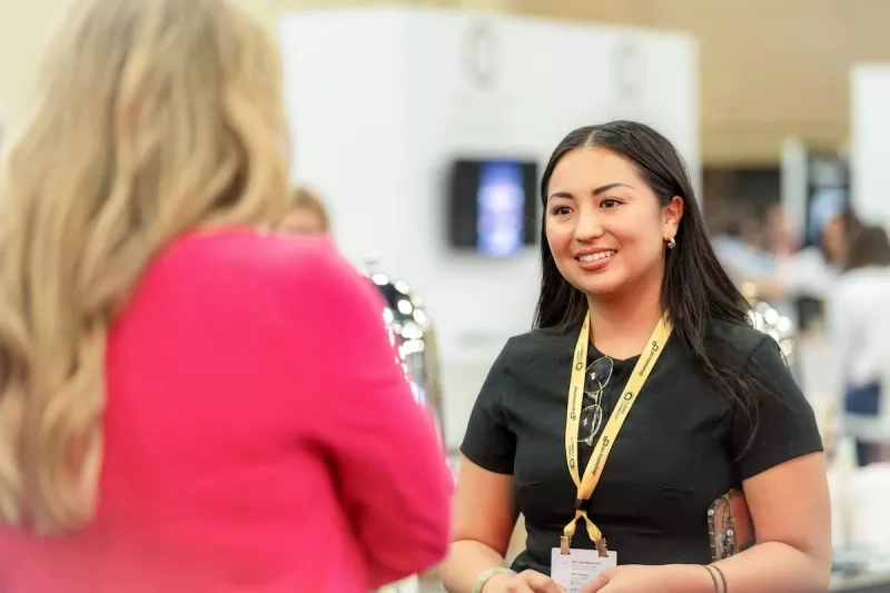 Woman in black shirt and yellow lanyard talking to woman in pink sweater at a trade show booth
