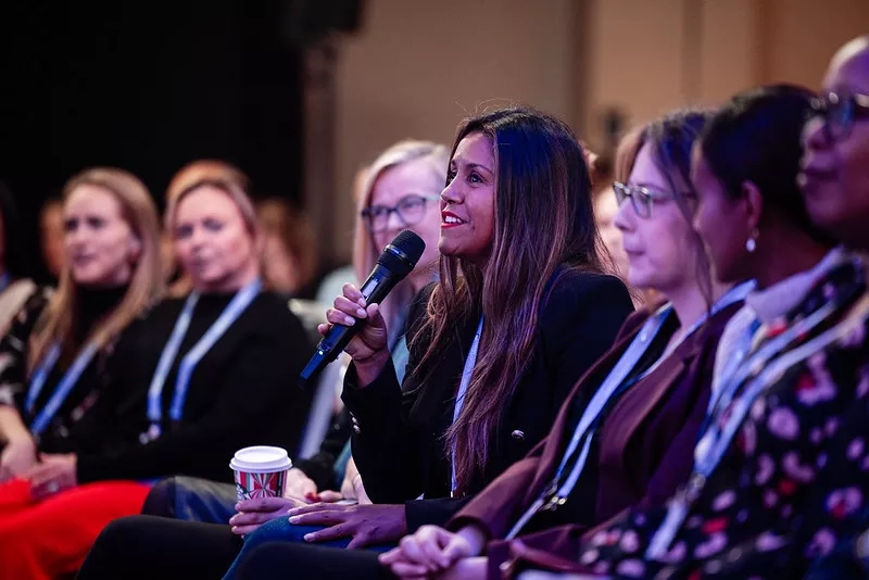 Picture of a lady holding a microphone asking a question sat in a conference audience