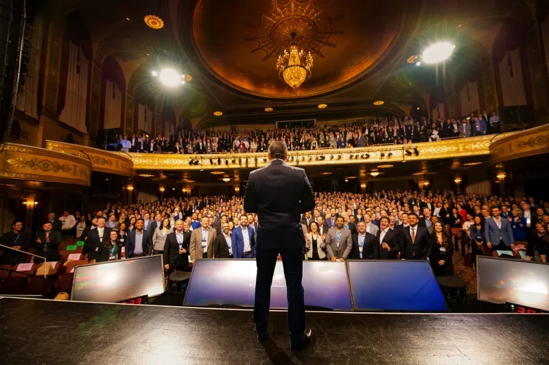 Speaker on stage facing a packed audience in an ornate theater at a large professional conference.