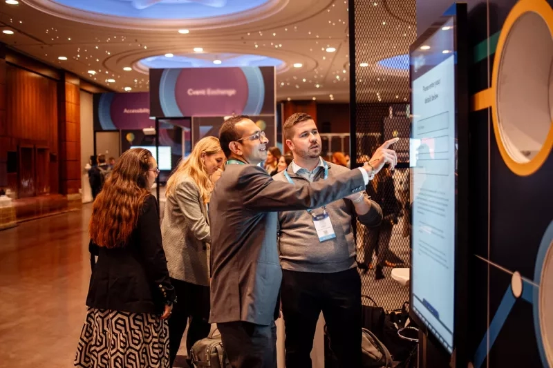 Group of conference attendees gathered around a large touchscreen display, one pointing at the screen.