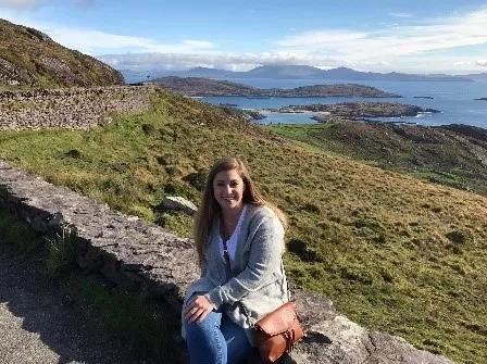 Michelle Williams sitting on a stone wall at the Cliffs of Moher with the sea in the background.