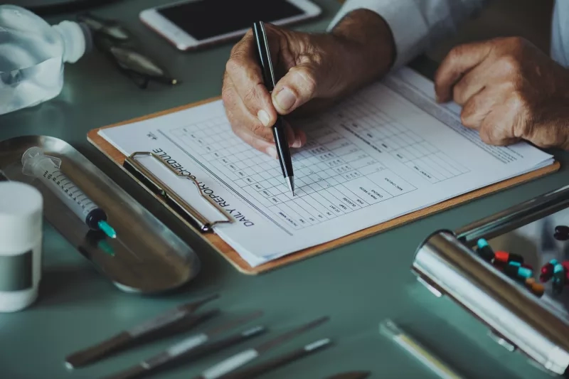 A man filling out a daily report with a clipboard and medical equipment on the table.