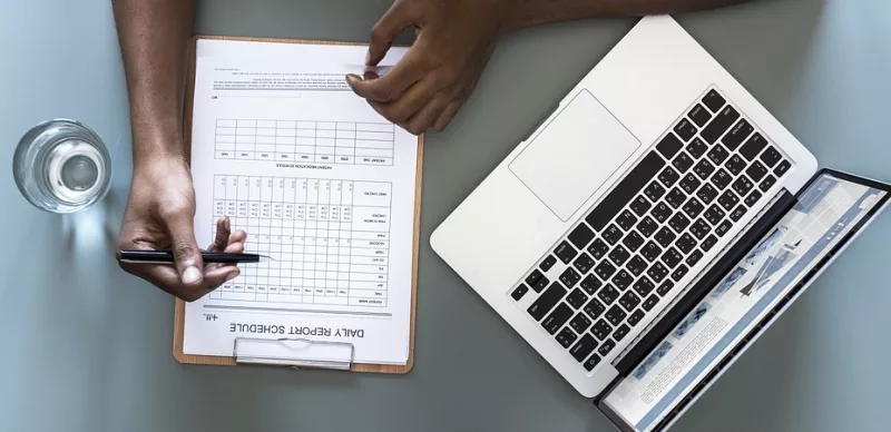 Hands holding pen over checklist, with laptop and water glass on gray table.