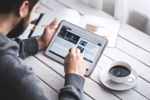 Man using tablet with cup of coffee on table, reading the news, and taking notes with a pen.