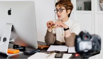 A woman with curly hair is sitting on a desk in front of an Apple computer.