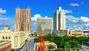 Downtown San Antonio with a blue sky, buildings, a red sculpture, trees, and a road with cars.