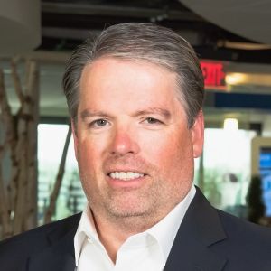 Close-up of a smiling man wearing a white shirt and black blazer with a tree in the background.
