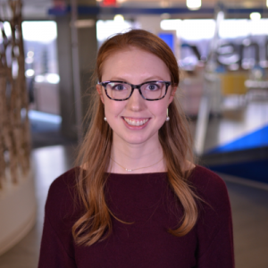 A woman with glasses and a maroon top is smiling in front of a bright building.