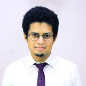 Portrait of a man with glasses, a white shirt, and a blue tie standing against a white wall.