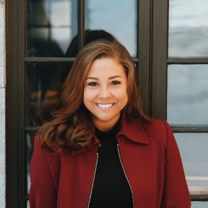 A smiling woman stands before a window, wearing a red jacket over a black shirt.