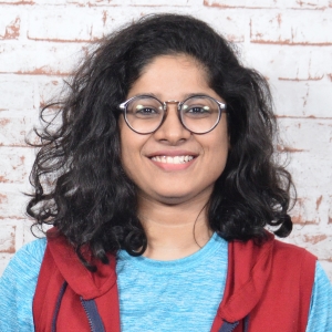 A lady wearing eyeglasses and a red vest smiling and posing for a picture against a brick wall.
