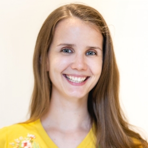A smiling woman in a yellow shirt with flower design in front of a white wall.