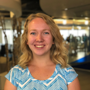 A smiling woman in a blue shirt stands in an indoor space with blurred background, possibly an office.